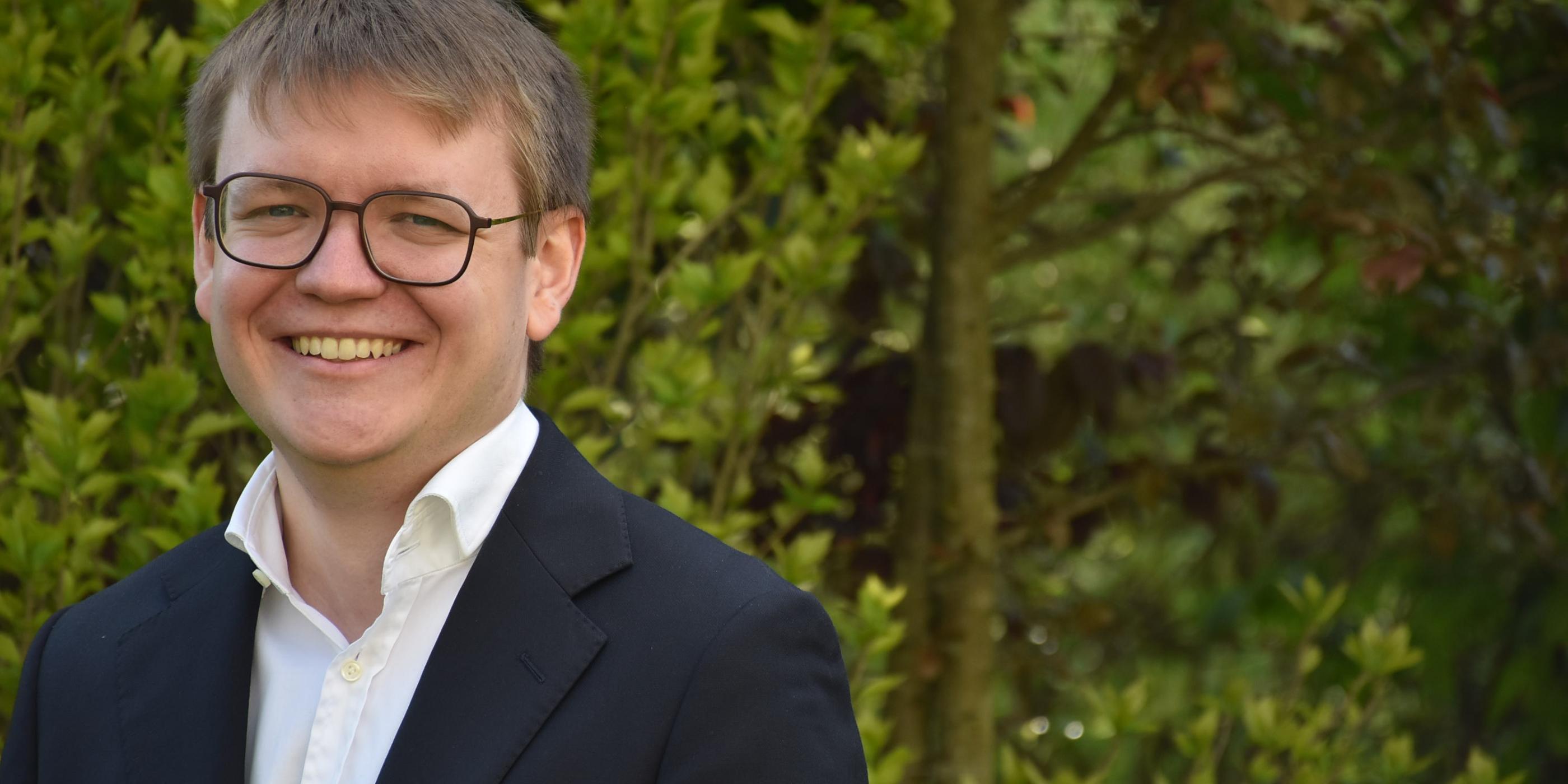 Portraitfoto von Pascal Klose. Im Hintergrund ein Busch mit grünen Blättern.