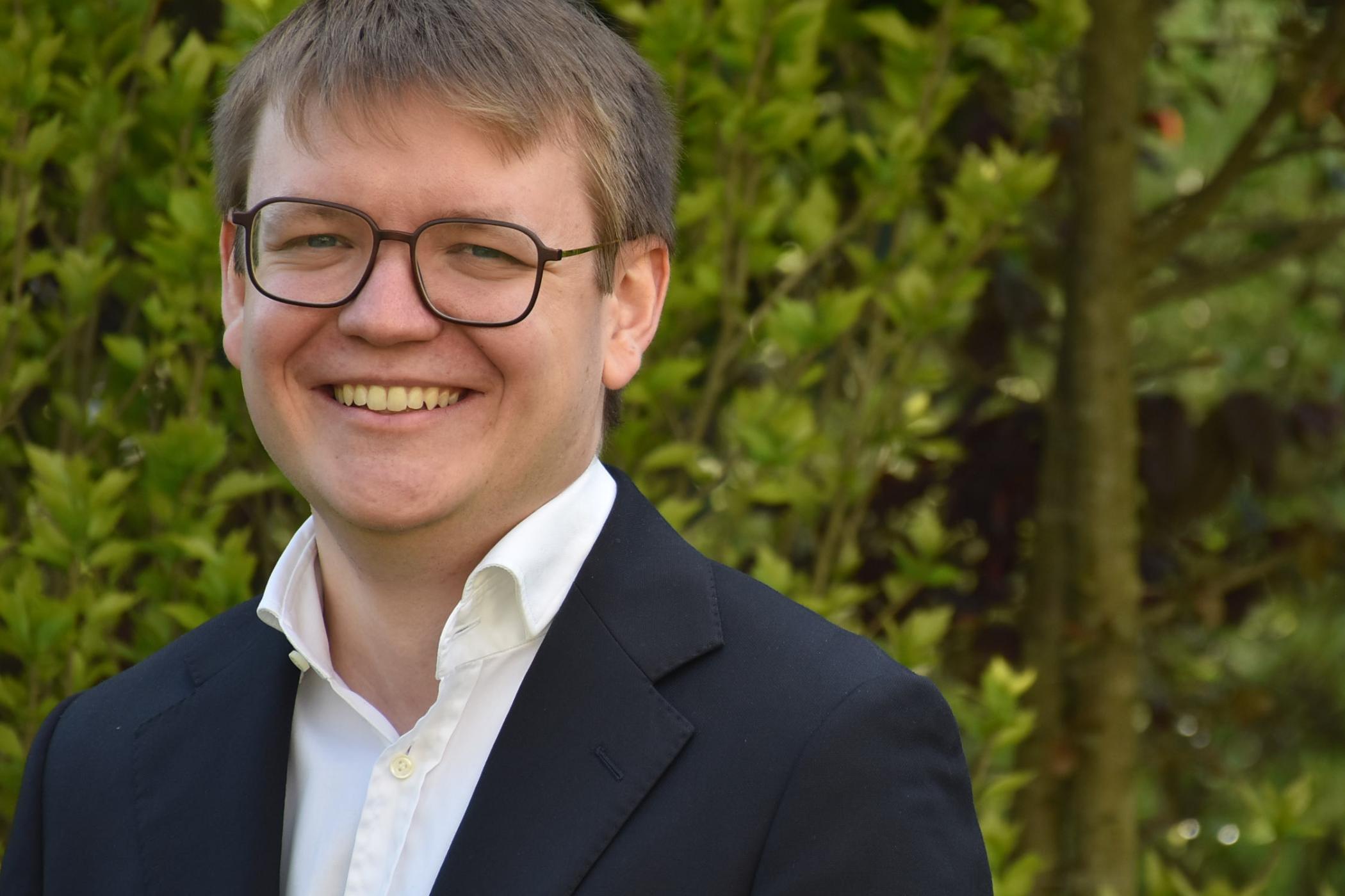 Portraitfoto von Pascal Klose. Im Hintergrund ein Busch mit grünen Blättern.