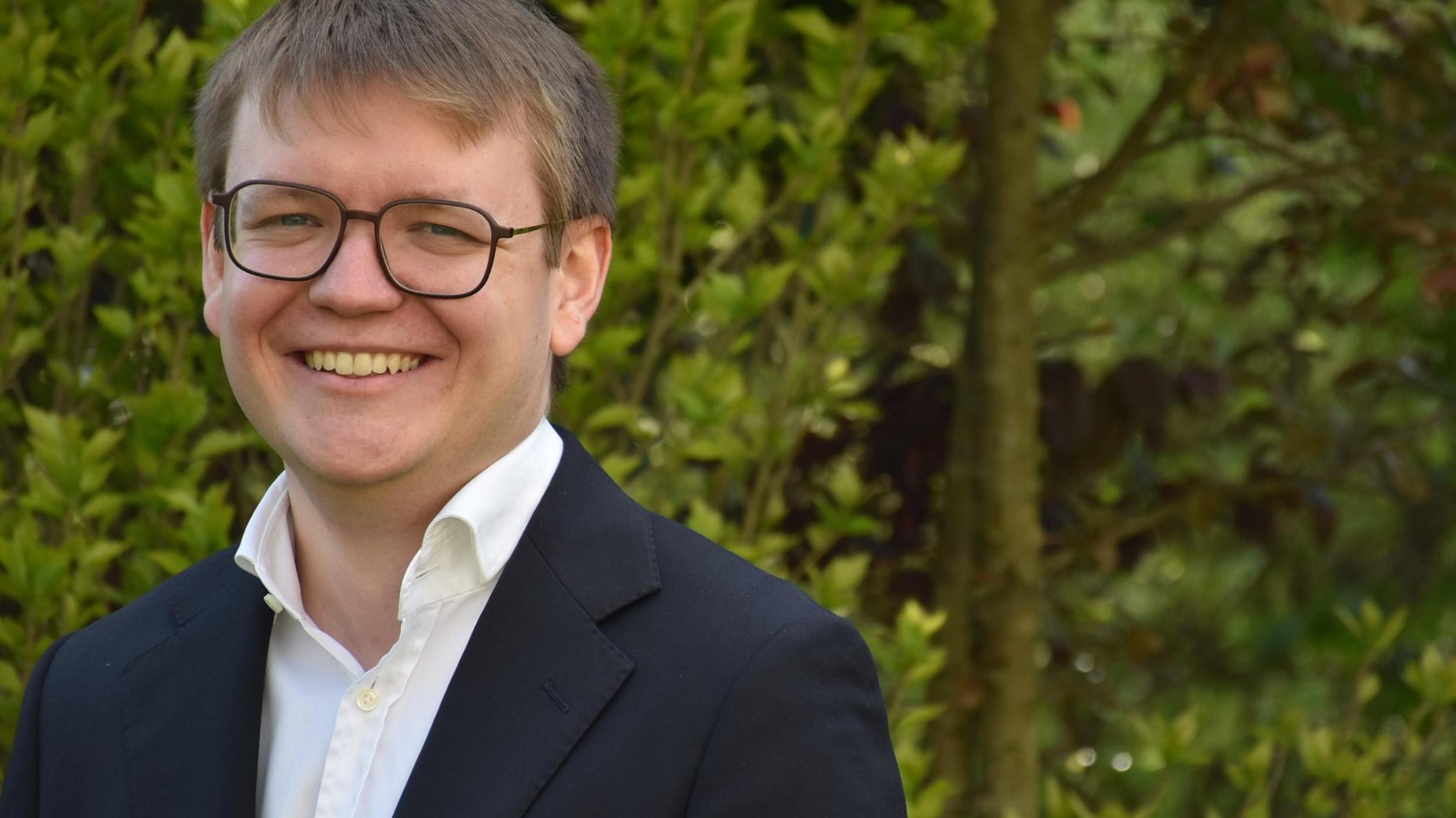 Portraitfoto von Pascal Klose. Im Hintergrund ein Busch mit grünen Blättern.