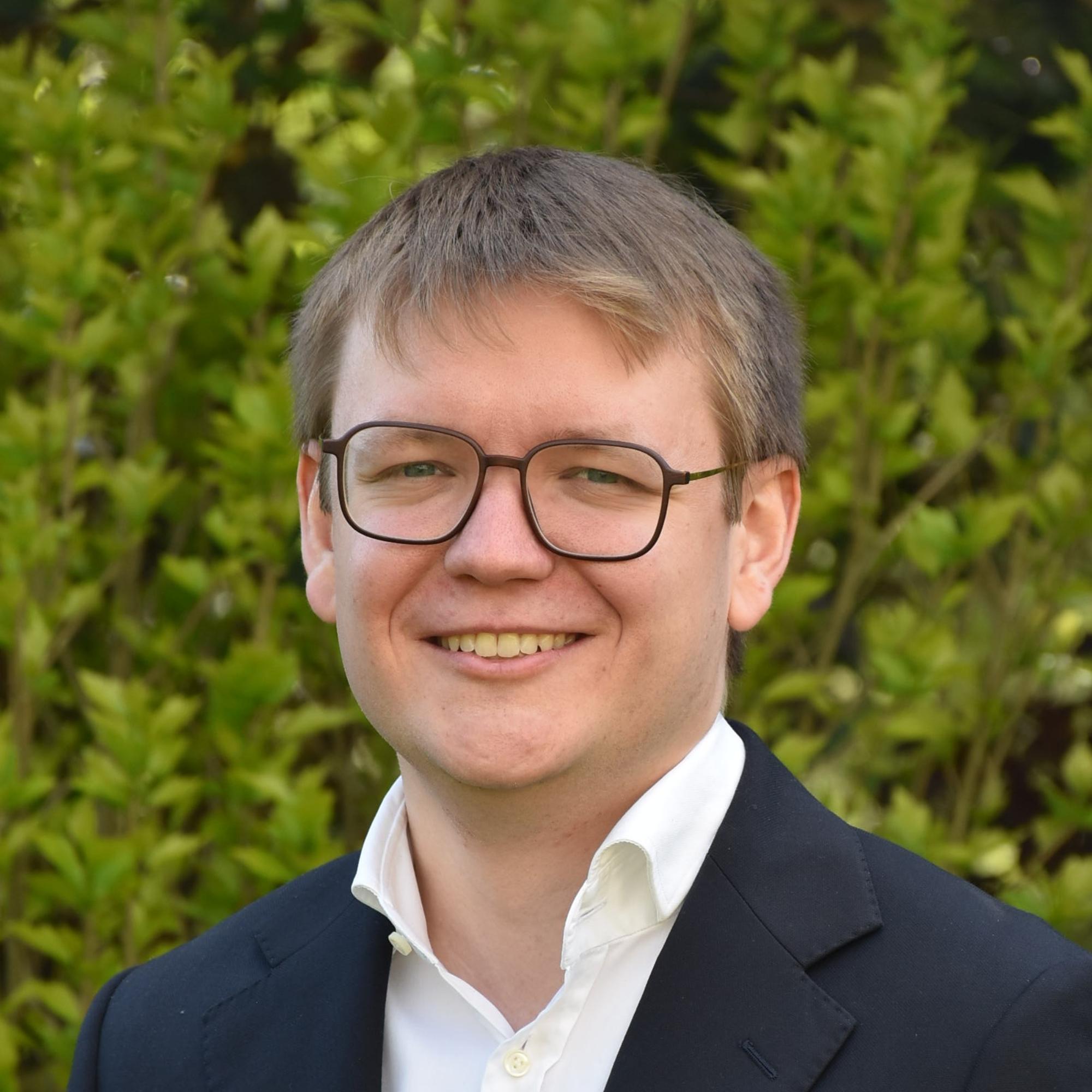 Portraitfoto von Pascal Klose. Im Hintergrund ein Busch mit grünen Blättern.