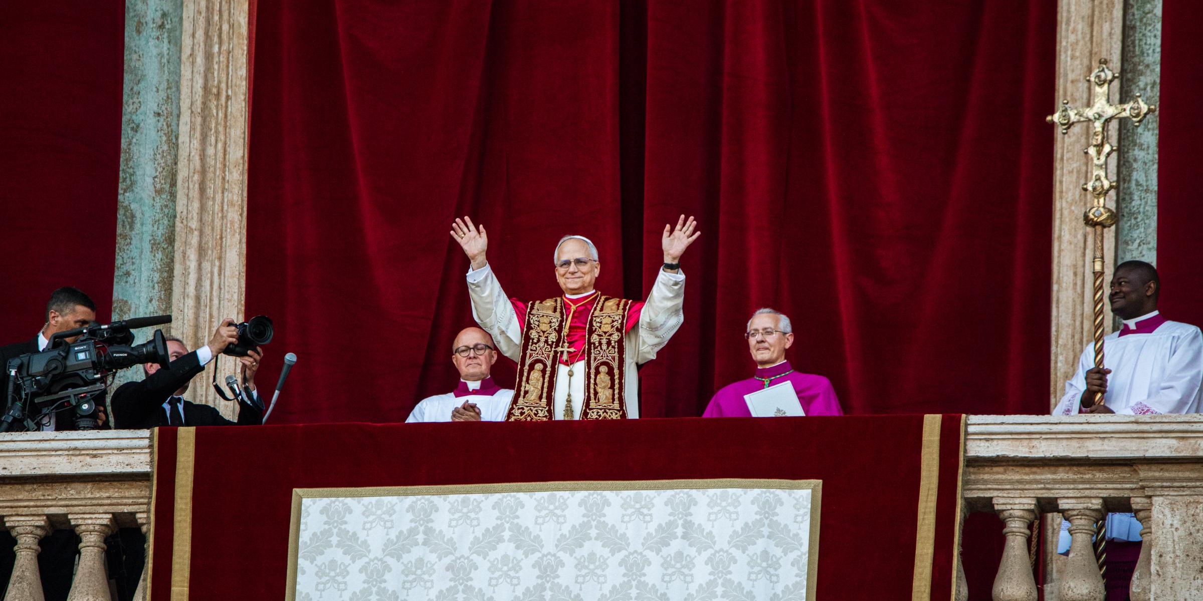 Der Papst steht auf dem Balkon des Petersdom und hebt beide Hände, zu seinem beiden Seiten sitzen zwei Kardinäle, hinter ihm ein roter Samtvorhang, an der Seite hängt ein Kreuz.